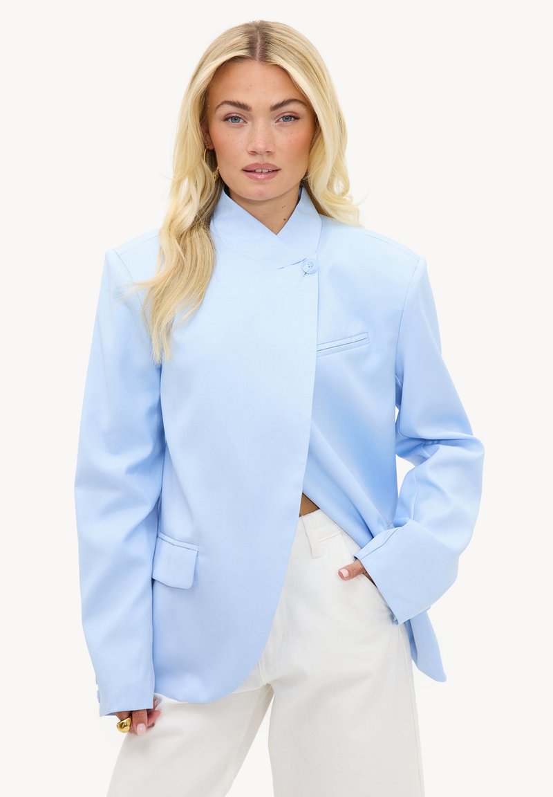 Femme blonde portant un blazer asymétrique bleu clair et un pantalon blanc, posant avec une main dans la poche sur fond blanc.