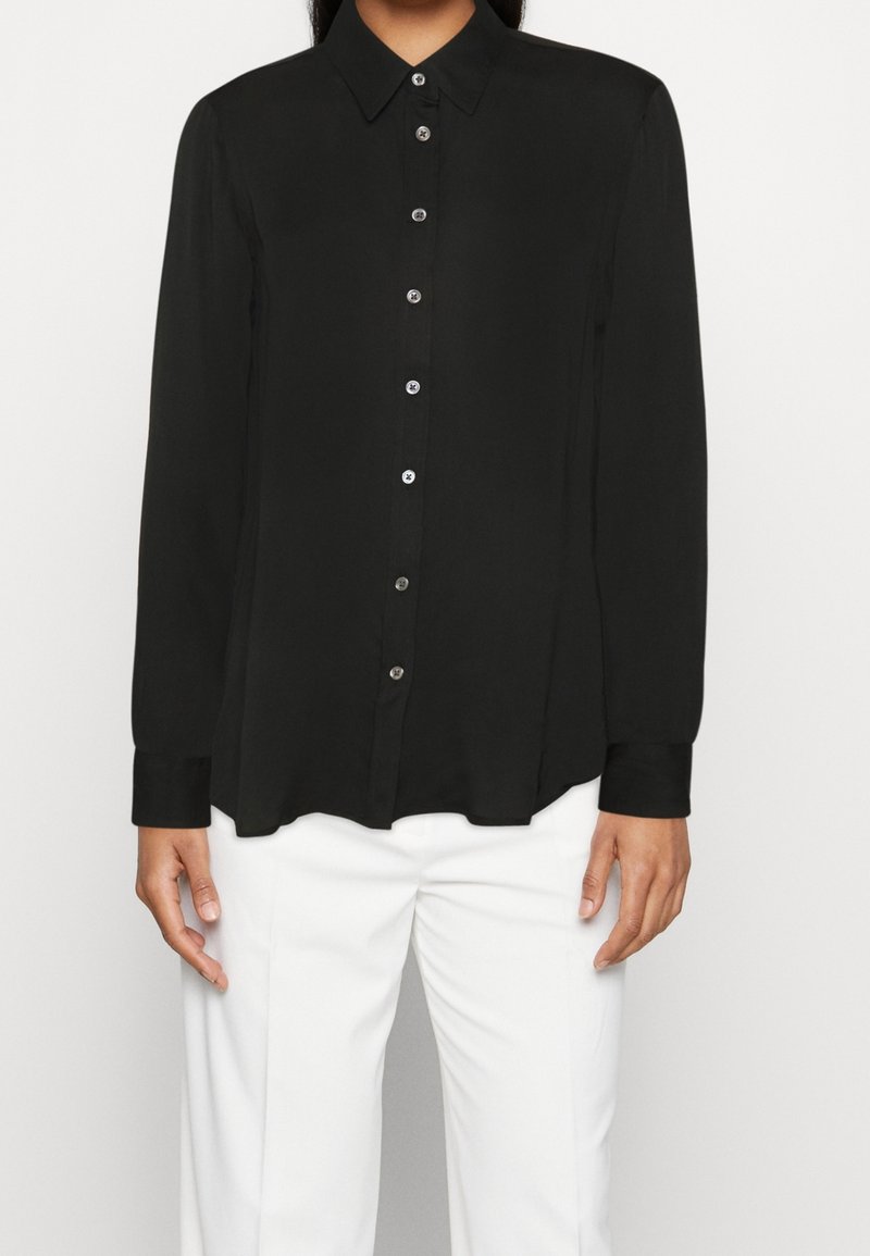 Chemise noire à manches longues avec un devant boutonné et un col classique. Fabriquée en tissu lisse, elle est dotée de boutons argentés et d'un ourlet légèrement arrondi.