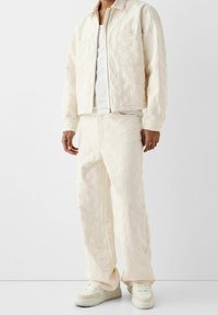 Ljus krämfärgad vadderad jacka med matchande byxor, prydd med fyrkantiga mönster och två framfickor, tillsammans med ljusa sneakers.