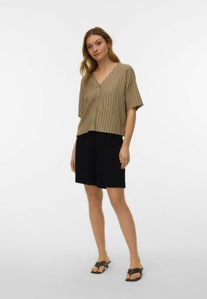 Kvinne som har på seg beige stripet kortermet V-hals skjorte med knapper, svarte shorts til knærne og mørke åpne sandaler, står foran hvit bakgrunn.