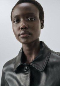 Zbliżenie na osobę z krótkimi kręconymi włosami, ubraną w czarną skórzaną kurtkę na jasnym, jednolitym tle.