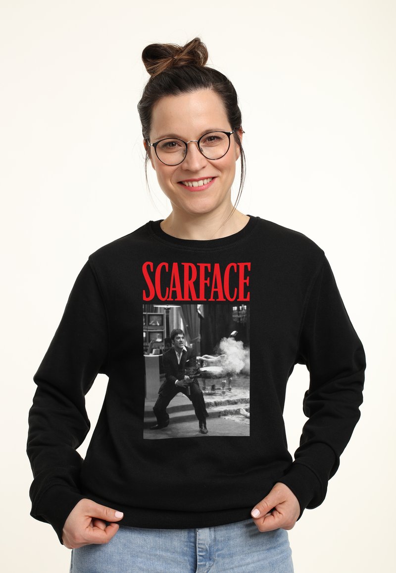 Felpa nera con un'immagine grande in bianco e nero di un personaggio con una pistola, incorniciata da testo rosso che legge "SCARFACE."