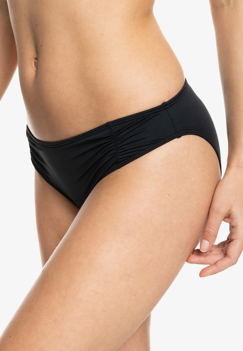 Melna bikini apakša ar sānu krokojumu, kas izceļ gludu tekstūru un vidēja augstuma dizainu, akcentējot vidukļa līniju.