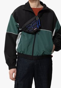 Persona que lleva una chaqueta a rayas en negro y verde oscuro con puños elásticos y una riñonera negra con detalles en cordones azules cruzando el pecho.