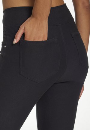 Eine Hand, die in die hintere Tasche einer schwarzen, figurbetonten Hose einer stehenden Person geschoben wird, zeigt die Taschendetails und die Textur des Stoffes.