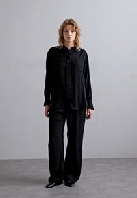 Cămașă neagră cu butoni, cu mâneci lungi, un buzunar pe piept, asortată cu pantaloni negri cu talie largă texturați și pantofi negri pe un fundal gri.