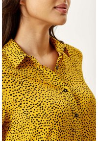 Yellow button-up shirt with black polka dots, featuring a collar and a smooth fabric texture. The shirt has a fitted design and dark buttons.