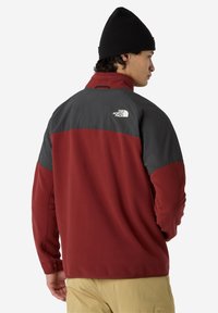 Hombre con un gorro negro y una chaqueta de forro polar roja y gris oscuro con el logo de The North Face en la parte superior de la espalda, de perfil.
