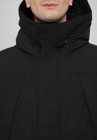 Jachetă neagră cu guler înalt și șnururi ajustabile, având o textură netedă și linii de design curate.