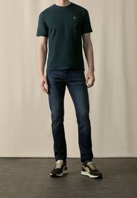 Man in een donkergroene t-shirt, slim-fit blauwe jeans en zwart-beige sneakers, staande tegen een lichtbeige achtergrond.