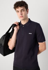 Ναυτικό πόλο μπλούζα με φερμουάρ στον γιακά και κοντά μανίκια, με λευκό λογότυπο στο στήθος και αντίθετη διακόσμηση στα μανίκια.