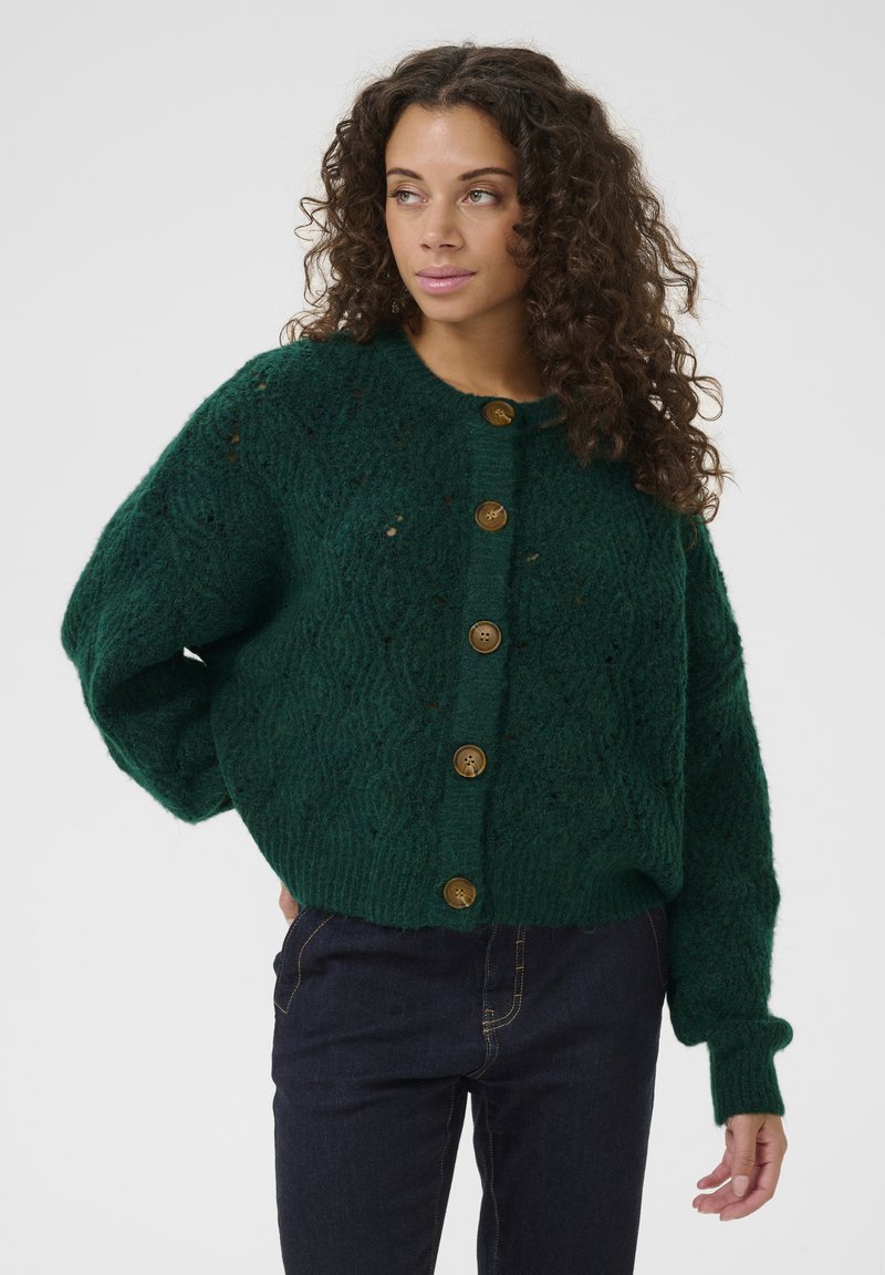 Zaļš adīts džemperis ar teksturētu rakstu, apaļu kaklu un sešām zelta krāsas pogām priekšpusē. Īss, brīvs piegriezums.