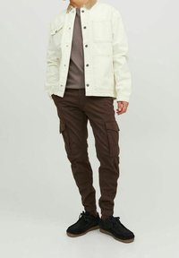 Giacca in denim crema con colletto marrone, camicia grigia sotto, pantaloni cargo marroni e sneakers nere con suola color cuoio.