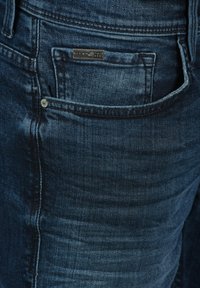 Tumma sininen farkut, joissa on klassinen viiden taskun muotoilu, pehmeä rakenne, hillitty kuluminen ja metallinen niitti etutaskussa.