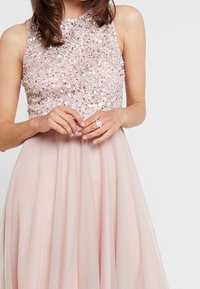 Bodice ozdobiony cekinami w delikatnym różowym kolorze, bez rękawów, z lekko opadającą przezroczystą tiulową spódnicą, tworzącą subtelną warstwową teksturę.