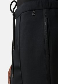 Zwarte joggingbroek met een gladde textuur, voorzien van een tailleband met trekkoord, zijzakken en een merkband langs de zijnaden.