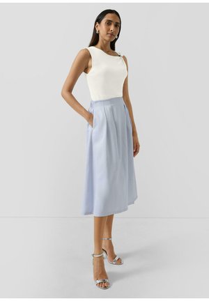 Kobieta stoi z ręką w kieszeni, ubrana w białą bluzkę bez rękawów, jasnoniebieską spódnicę midi oraz srebrne szpilki z paskiem wokół kostki na szarym tle.