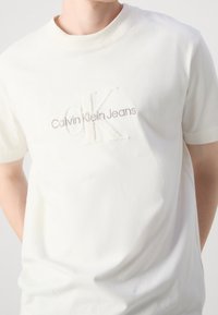Krémové bavlněné tričko s kulatým výstřihem. Obsahuje reliéfní logo "CK" a text "Calvin Klein Jeans" v kontrastní béžové barvě.