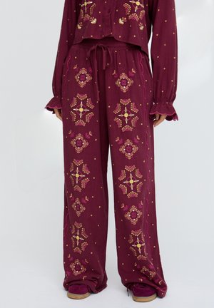 Weinrote Hosen mit weitem Bein, gelber und rosa geometrischer Stickerei, Kordelzug in der Taille, getragen mit passendem Langarmshirt und weinroten Schuhen.