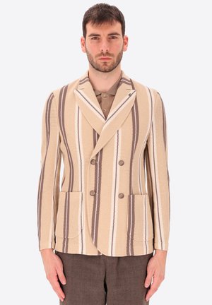 Uomo che indossa un blazer doppiopetto beige e marrone a righe verticali su una camicia marrone e pantaloni scuri, in piedi davanti a uno sfondo semplice.