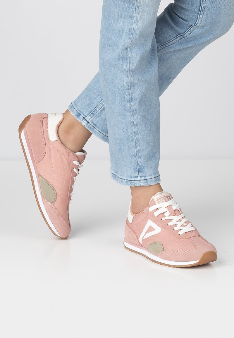 Personne portant un jean bleu clair et des baskets roses avec des lacets blancs et des accents beiges, debout sur un sol blanc devant un fond uni.