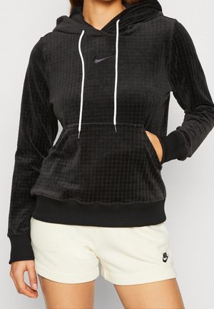 Kobieta w czarnej, teksturowanej bluzie z kapturem Nike z białymi sznurkami i białych szortach Nike z logo na lewej nodze.