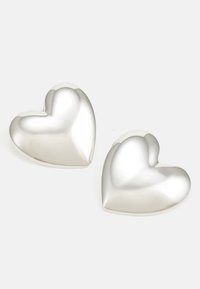 Dos piezas decorativas de plata en forma de corazón con una textura suave y brillante y bordes redondeados, reflejando la luz sobre un fondo blanco.