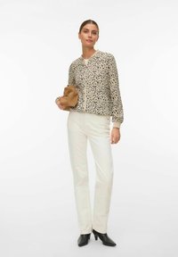 Sieviete valkā leoparddrukas rāvējslēdzēja jakas, baltas bikses, melnus augstpapēžu zābakus un tur brūnu cepuri uz baltas fona.