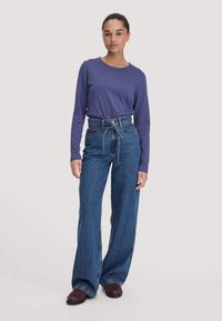 Lila Langarmoberteil kombiniert mit hoch taillierten blauen weiten Jeans, die einen gebundenen Gürtel und eine lockere Passform aufweisen, ergänzt durch dunkle Schuhe.