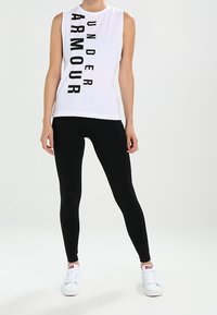 Haut blanc sans manches avec texte noir "Under Armour" à l'avant, associé à des leggings noirs et des baskets blanches. Design simple et ajusté.