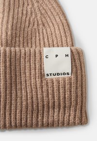 Lichtbruin geribd gebreide beanie met een wit label waarop "CPH STUDIOS" in zwarte letters staat. Zachte textuur en een opgevouwen manchet-detail.