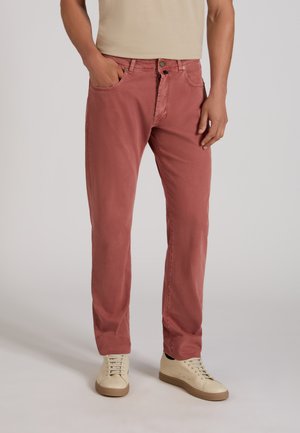 Mężczyzna w wyblakłych czerwonych spodniach, beżowej koszuli i kremowych trampkach, stojący z lewą ręką w kieszeni na jednolitym tle.