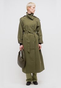 Άτομο με κοντά ξανθά μαλλιά που φοράει ένα χακί παλτό με ζώνη, χακί παντελόνι, μαύρα παπούτσια, κρατώντας μια υφασμάτινη χακί τσάντα, κοιτάζοντας προς τα δεξιά.