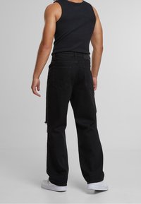 Homme portant un débardeur noir, un jean noir ample avec poches arrière et des baskets blanches, debout dos à la caméra.