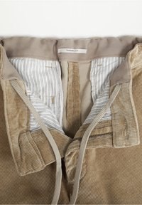 Pantalon en velours côtelé beige avec une fermeture éclair ouverte et un cordon, montrant une doublure en tissu rayé et une étiquette « Tapered Fit » à l'intérieur de la ceinture.