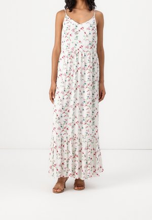 Femme portant une robe maxi blanche sans manches avec des fines bretelles et un petit motif floral rouge et bleu, associée à des sandales marron.