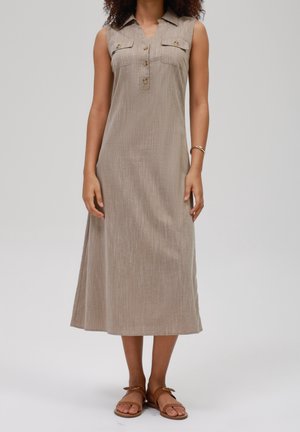 Vrouw draagt een mouwloze beige midi-jurk met knoopsluitingen op de borstzakken en bruine platte sandalen, staand tegen een effen achtergrond.