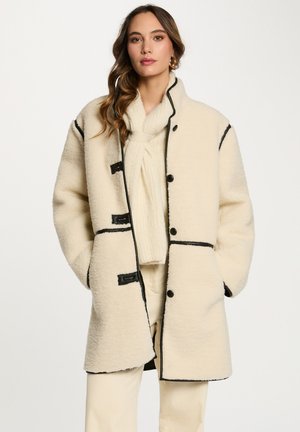 Cappotto in pile color crema con rifiniture nere, caratterizzato da un collo alto e bottoni frontali. Texture morbida con una vestibilità rilassata e over-size.