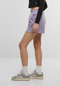 Shorts di cotone lilla con vita elastica, tasche laterali e design con ricamo di farfalle. Indossati con sneakers grigie e calzini bianchi.