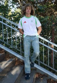 Maglia sportiva bianca con accenti verdi, logo Adidas ed emblema; abbinata a jeans azzurri chiari e sneaker nere, posata su scale.