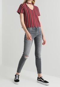 Top de rayas rojas y blancas de manga corta con escote en V y detalle de choker, combinado con jeans ajustados grises que presentan acentos desgastados y zapatillas negras.