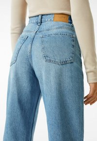 Person som bär ljusblå högmidjade jeans och en beige långärmad topp, visad bakifrån från midjan ner till mitten av låret.