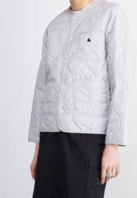 Persona vistiendo una chaqueta acolchada gris claro con botones a presión, bolsillo en el pecho con logo, y pantalones negros con bolsillo lateral, mano con anillo de plata.