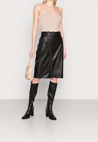 Svart läder midi kjol med dragkedja i fronten, bälte i midjan, kombinerad med knähöga svarta stövlar och en beige ribbad bodysuit samt en handväska.