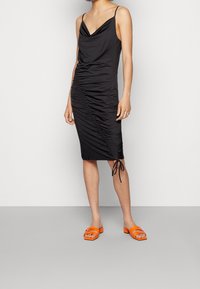 Žena v bezramínkových černých midi šatech s nařasením a bočním uzlem, v kombinaci s jasně oranžovými sandály na klínku, stojí na šedé podlaze a u bílé zdi.