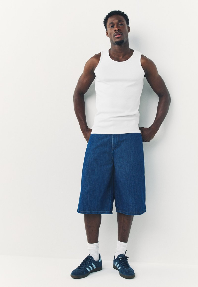 Jeune homme portant un débardeur blanc, un short en jean long, des chaussettes blanches et des baskets bleu marine, debout contre un mur blanc.