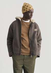 Άνδρας που φοράει καφέ μπουφάν με επένδυση από shearling, καφέ φούτερ, ελαιδί παντελόνι και κίτρινο σκουφάκι με καφέ πουά, στέκεται με τα χέρια στις τσέπες.