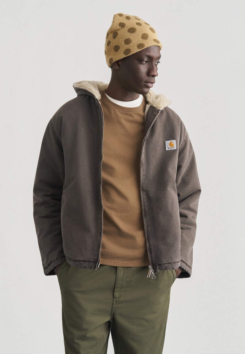 Mann trägt eine braune Jacke mit Schaffellfutter, einen beigefarbenen Pullover, olivgrüne Hosen und eine gelbe Mütze mit braunen Punkten, steht mit den Händen in den Taschen.