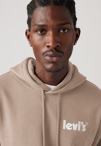 Beige hoodie gemaakt van zacht materiaal, met een voorzak en een witte "levi's" logo in glanzende print op de linker borst.