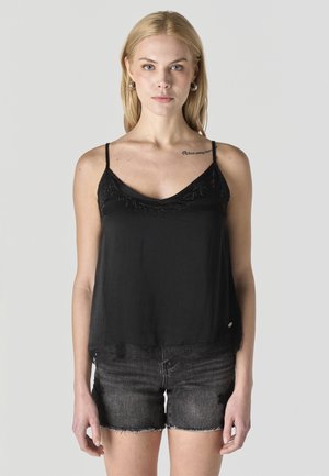 Mujer rubia con pendientes de aro que lleva un top negro sin mangas y pantalones cortos de mezclilla oscuros desgastados sobre un fondo gris liso.